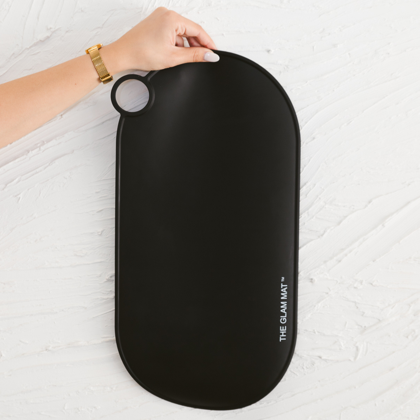 A black, oval-shaped silicone mat with a small round handle at one end, branded with 'THE GLAM MAT'.